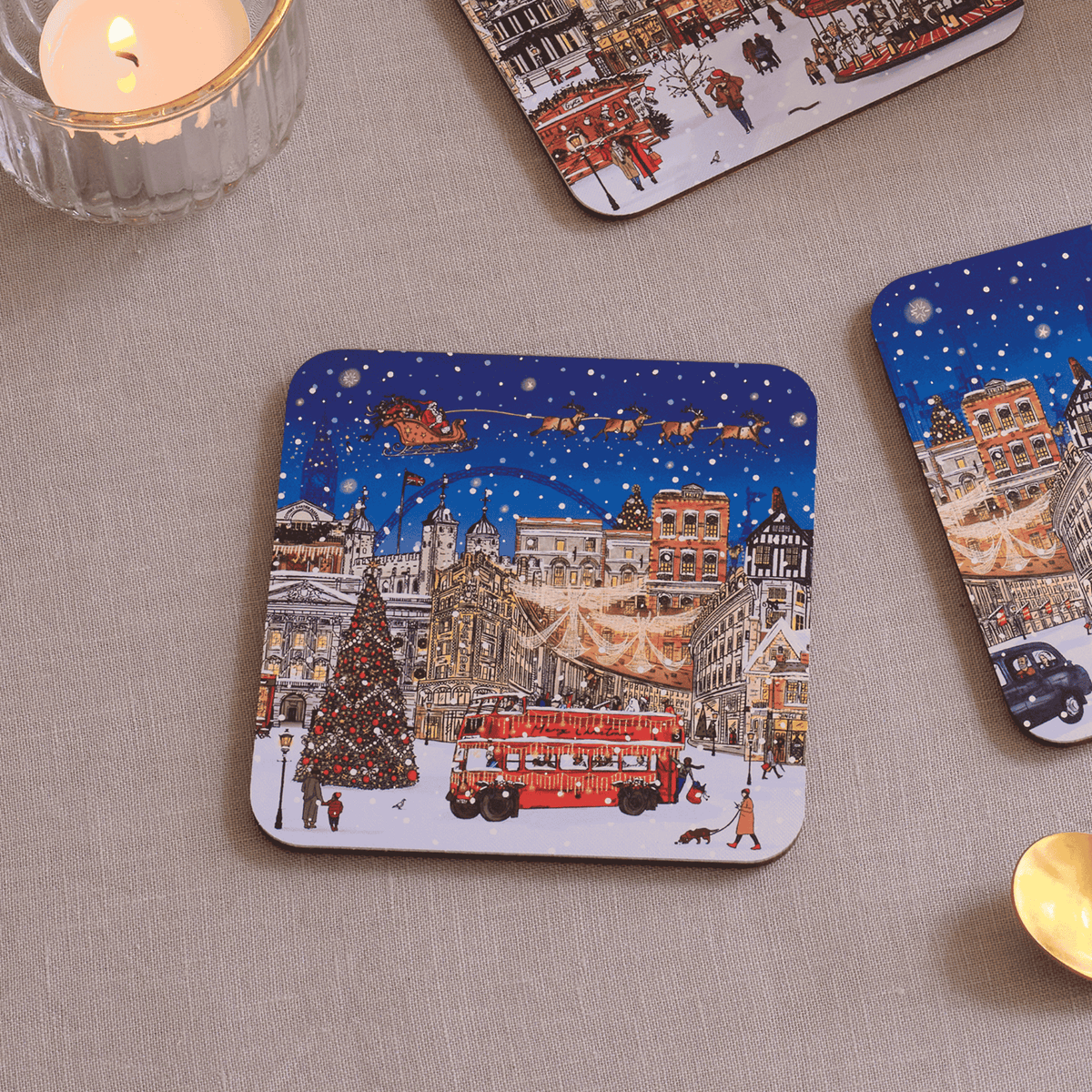 A square coaster with rounded corners featuring a festive Christmas city scene, displayed on a beige fabric surface alongside other coasters and lit candles.