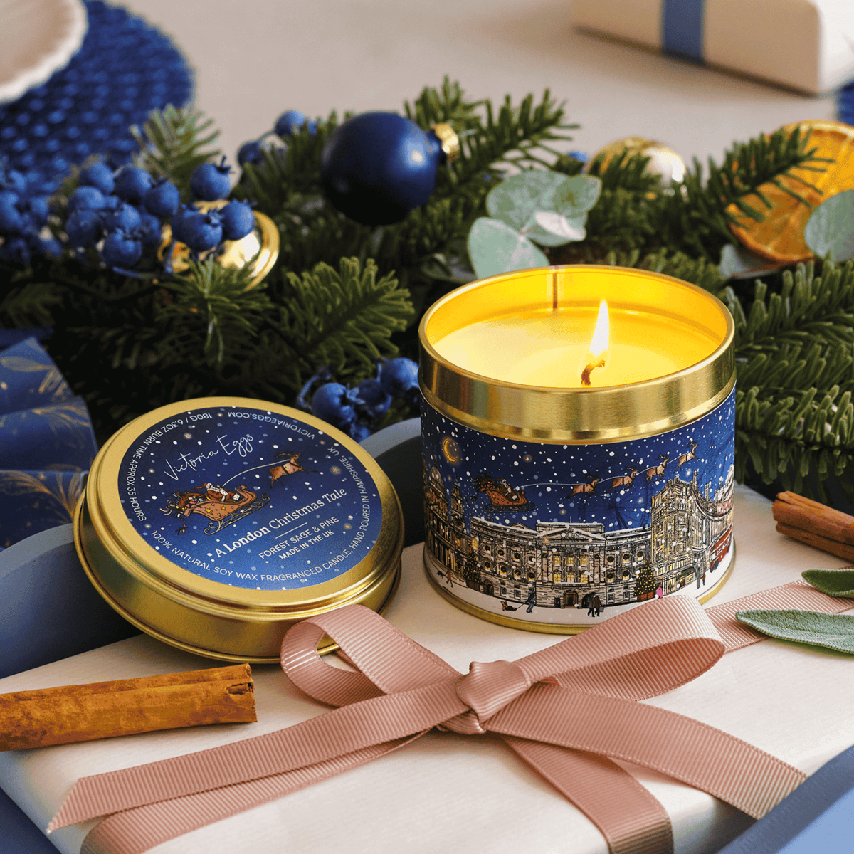 A lit cylindrical candle in a gold-rimmed tin with a winter cityscape label, placed beside its matching lid on a wrapped gift. The scene is styled with greenery, blue ornaments, cinnamon sticks, and a pink ribbon.
