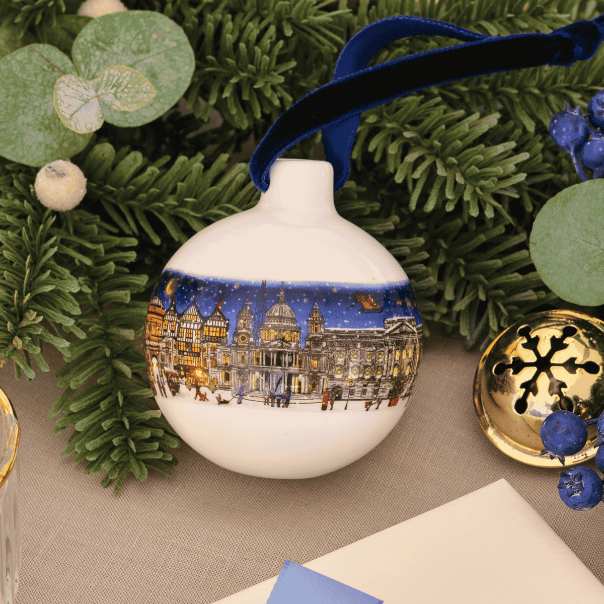 A round white Christmas ornament featuring a detailed winter night scene of London's St. Paul's Cathedral and surrounding street, hanging from a dark blue ribbon.