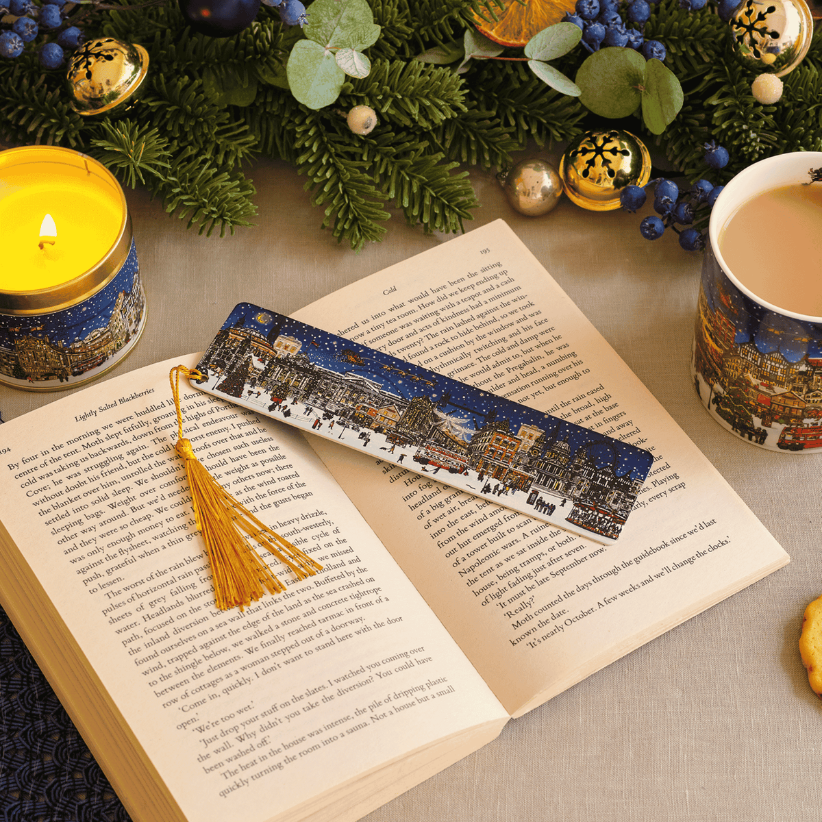 A decorative book bookmark with a tassel lies across an open book on a tabletop. The bookmark features a printed winter cityscape scene in blue, white, and warm accent tones.