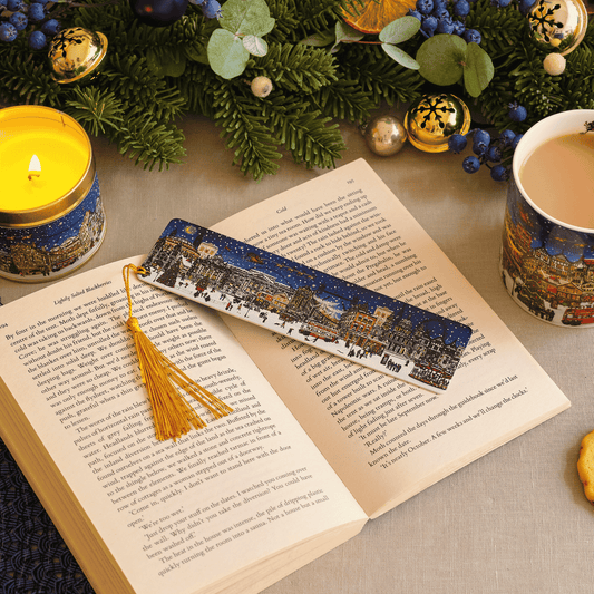 A decorative book bookmark with a tassel lies across an open book on a tabletop. The bookmark features a printed winter cityscape scene in blue, white, and warm accent tones.