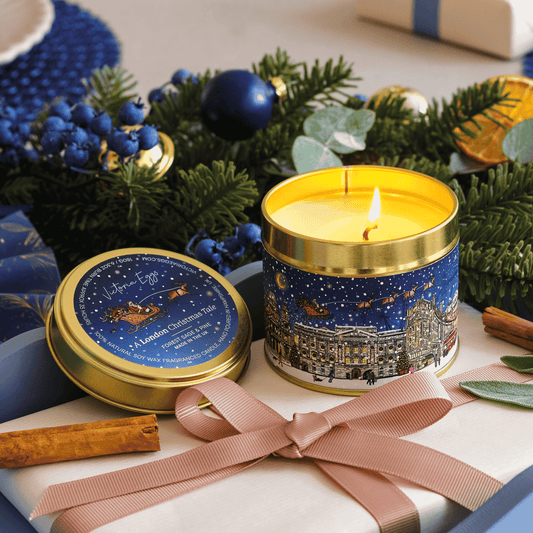 A lit cylindrical candle in a gold-rimmed tin with a winter cityscape label, placed beside its matching lid on a wrapped gift. The scene is styled with greenery, blue ornaments, cinnamon sticks, and a pink ribbon.