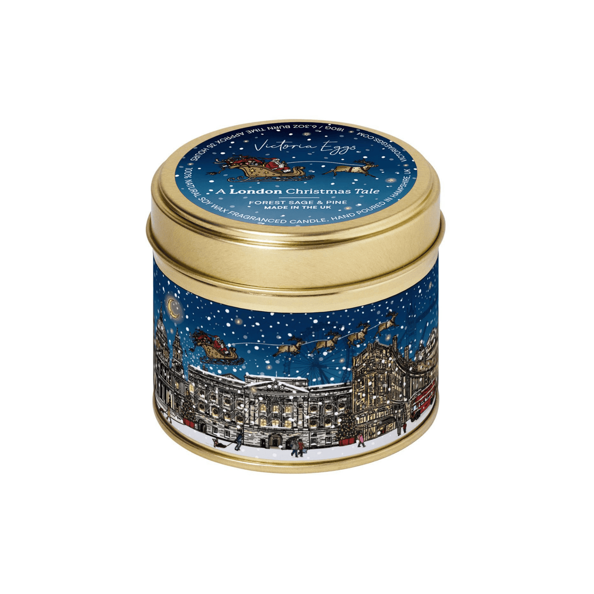 A round metal candle tin with a gold lid and a blue illustrated label depicting a snowy London street scene at night, photographed on a plain white background.